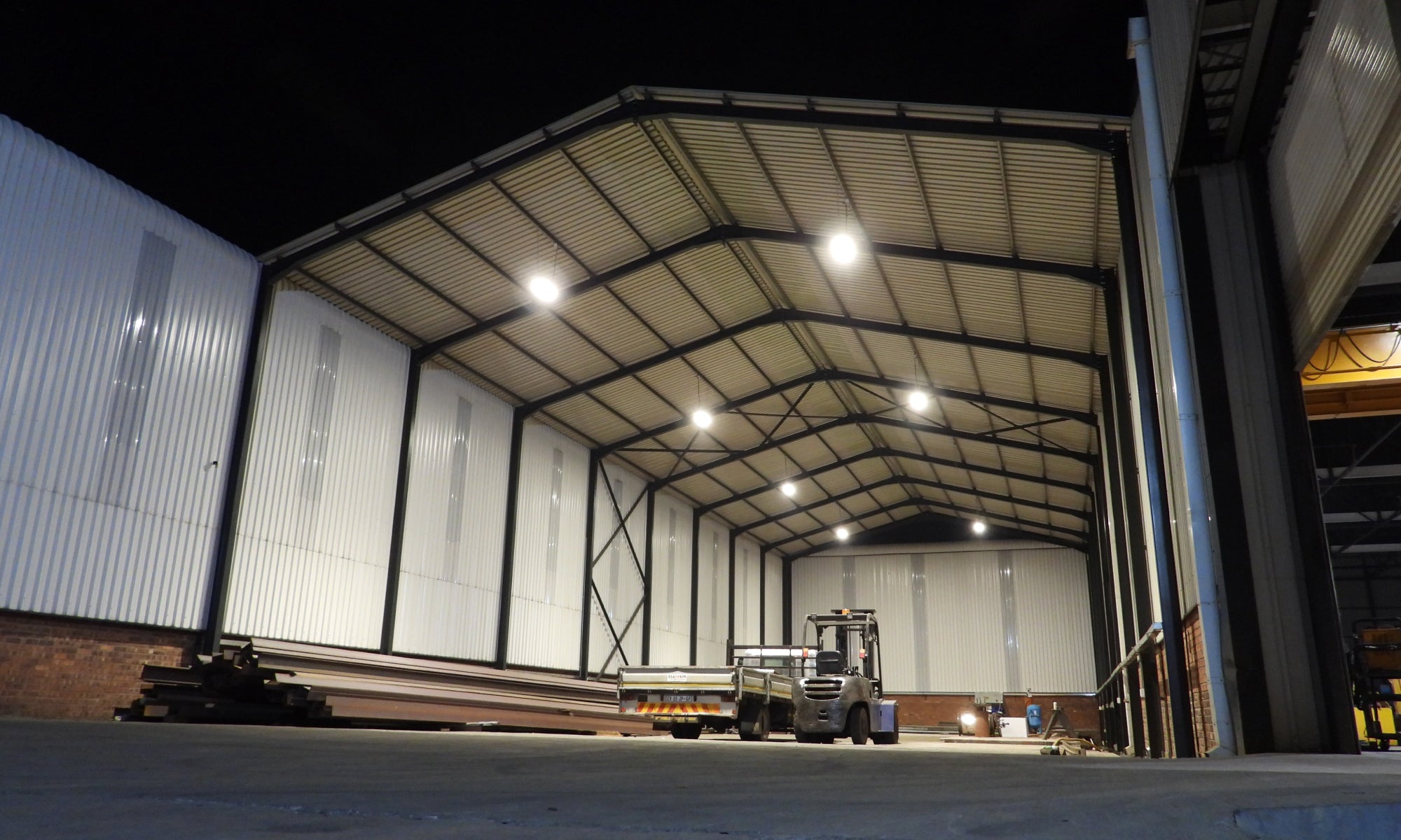 Large industrial warehouse with metal beams and a forklift at night, showcasing effective Commercial Lighting solutions.